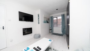 Une chambre moderne avec un mur blanc, un téléviseur à écran plat fixé au-dessus d’une table blanche, une fenêtre avec des rideaux et une porte.