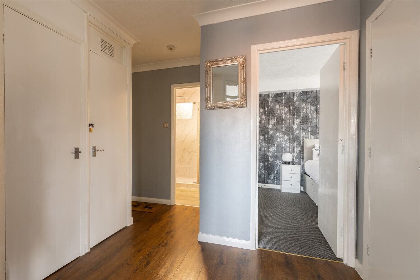 A hallway with wooden flooring leading to a bedroom with a bed, a nightstand, and a mirror on the wall.