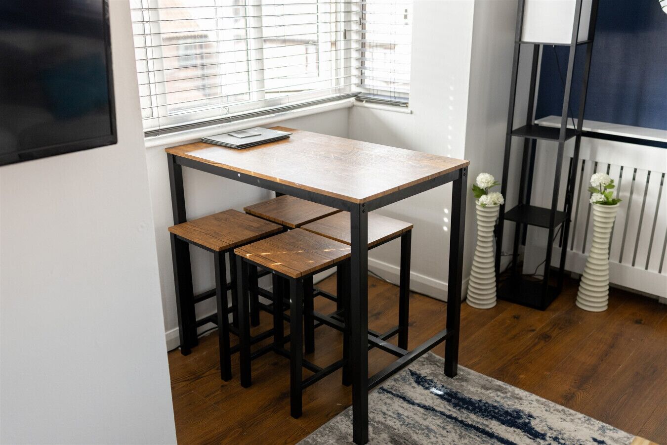 A small, modern kitchen with a wooden table and stools, a window with blinds, and a radiator.