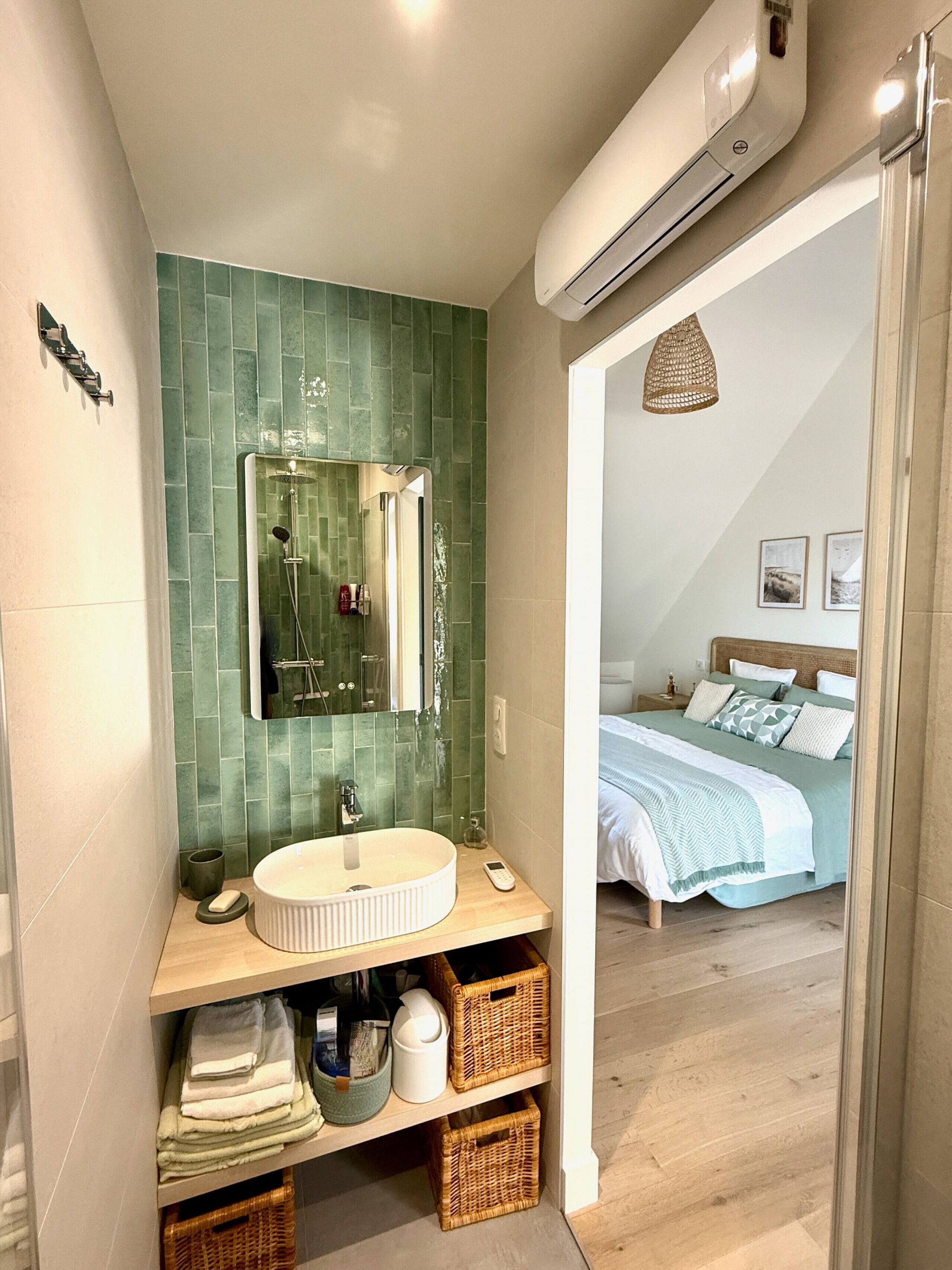 A bathroom with a green-tiled wall, a round sink, a mirror, a shower, and a bedside table with a lamp.