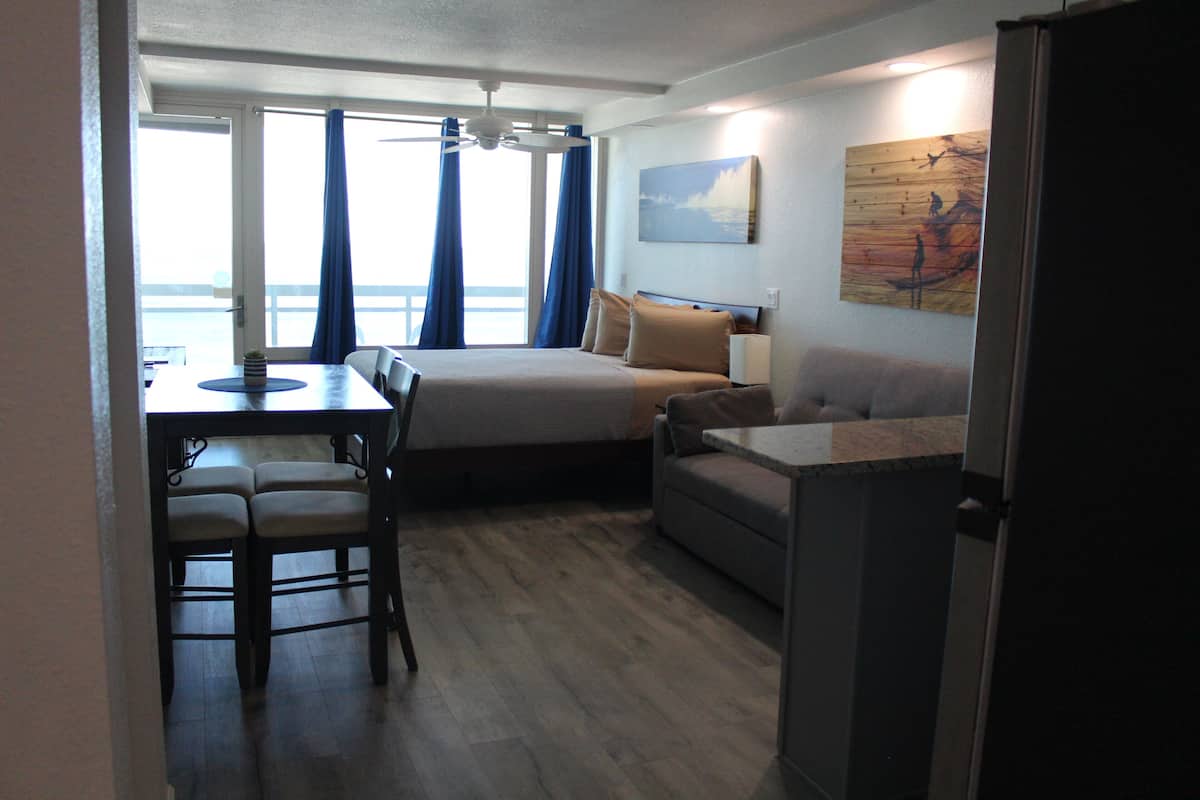 A compact hotel room with a bed, a sofa, a dining table with chairs, a small kitchen area, and a large window with curtains.