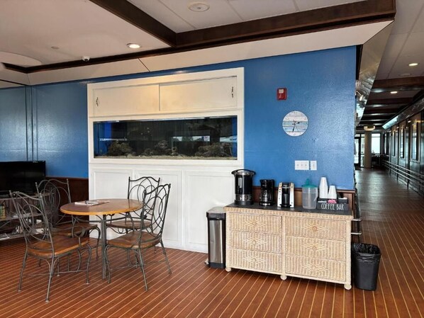 A room with a blue wall, a white cabinet, a round table with chairs, a coffee bar, and a trash bin. - Georgian Inn Beach Club (Ormond Beach)
