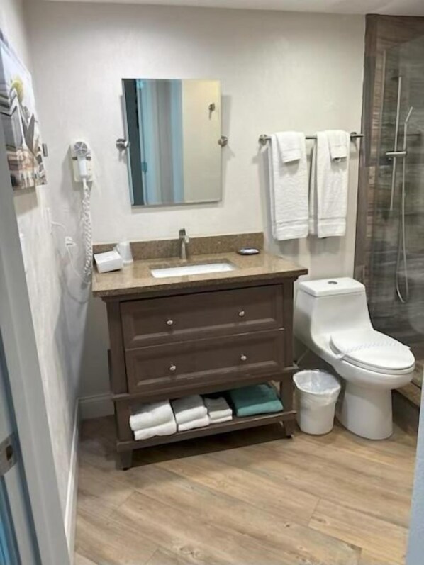 A bathroom with a wooden vanity, a sink, a mirror, a toilet, and a shower area. - Georgian Inn Beach Club (Ormond Beach)