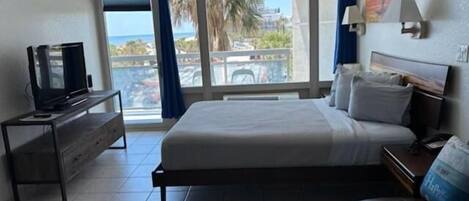 Een slaapkamer met een bed, een televisie, een bank en een balkon met uitzicht op palmbomen en de zee.