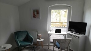Ein kleiner, gut beleuchteter Raum mit einem türkisfarbenen Sessel, einem weißen Schreibtisch mit einem Laptop, einem Fernseher und einem Korbstuhl.