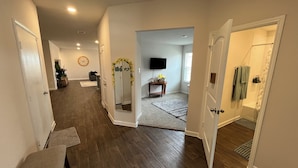 A hallway with wooden flooring, white walls, and doors leading to a bathroom and a living area with a TV.