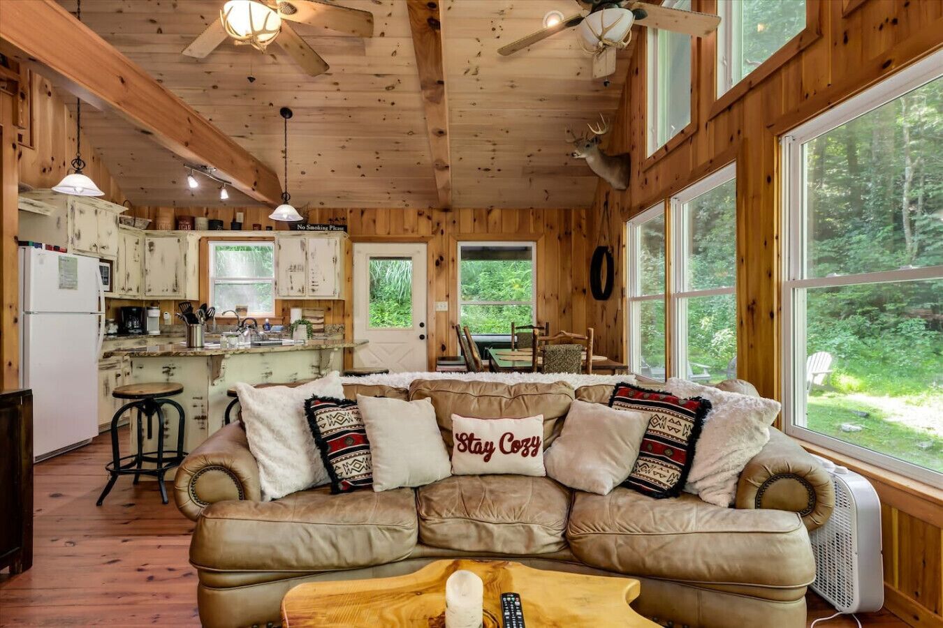 A cozy cabin interior with a wooden ceiling, a kitchen area, a large window, and a leather sofa with decorative pillows.