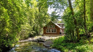 A cabin with a wooden exterior, surrounded by dense forest and a flowing stream.
