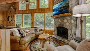 A cozy cabin interior with a stone fireplace, a television mounted above it, and a large window offering a view of the surrounding forest.