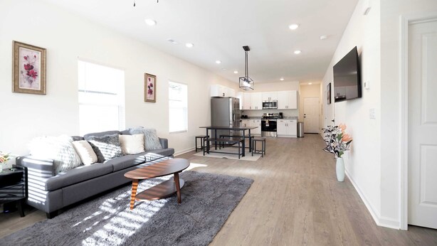 A modern living room with a grey sofa, a coffee table, and a rug. A kitchen with white cabinets and a dining area is visible in the background. - 4 Min to Downtown | 10 to Elon | Car Rental Avail. (Burlington)
