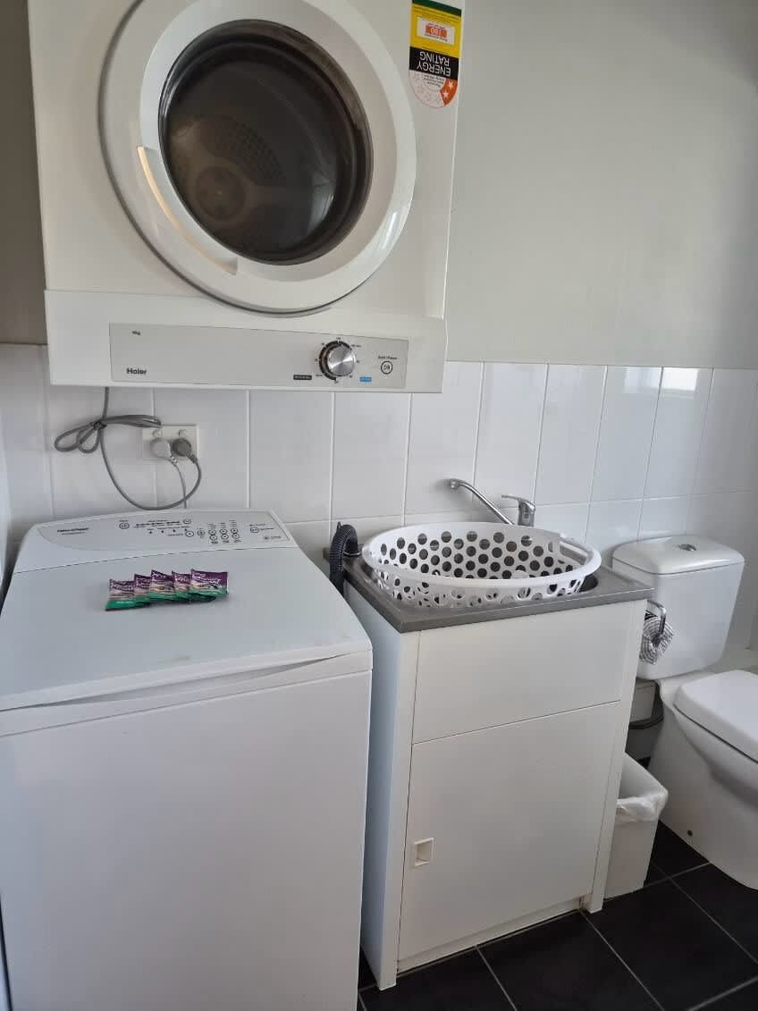 Uma lavanderia compacta com máquina de lavar frontal, uma pia pequena com escorredor e vaso sanitário.