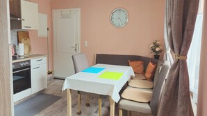 Ein kompakter Wohnraum mit Küchenzeile, einem Esstisch mit Stühlen, einer Couch mit Kissen und einer Uhr an der Wand.