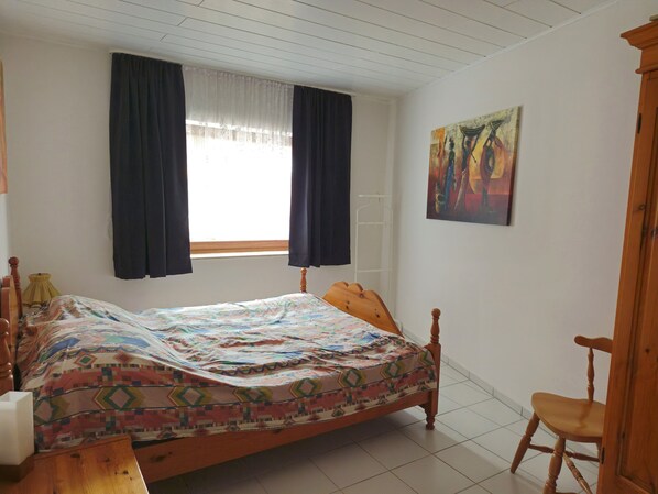 Un dormitorio con cama, una silla, una ventana con cortinas, un cuadro en la pared y un techo blanco.
