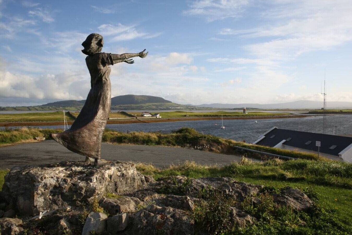 A statue of a figure with outstretched arms on a rock by a body of water with a view of hills and a lighthouse in the distance.