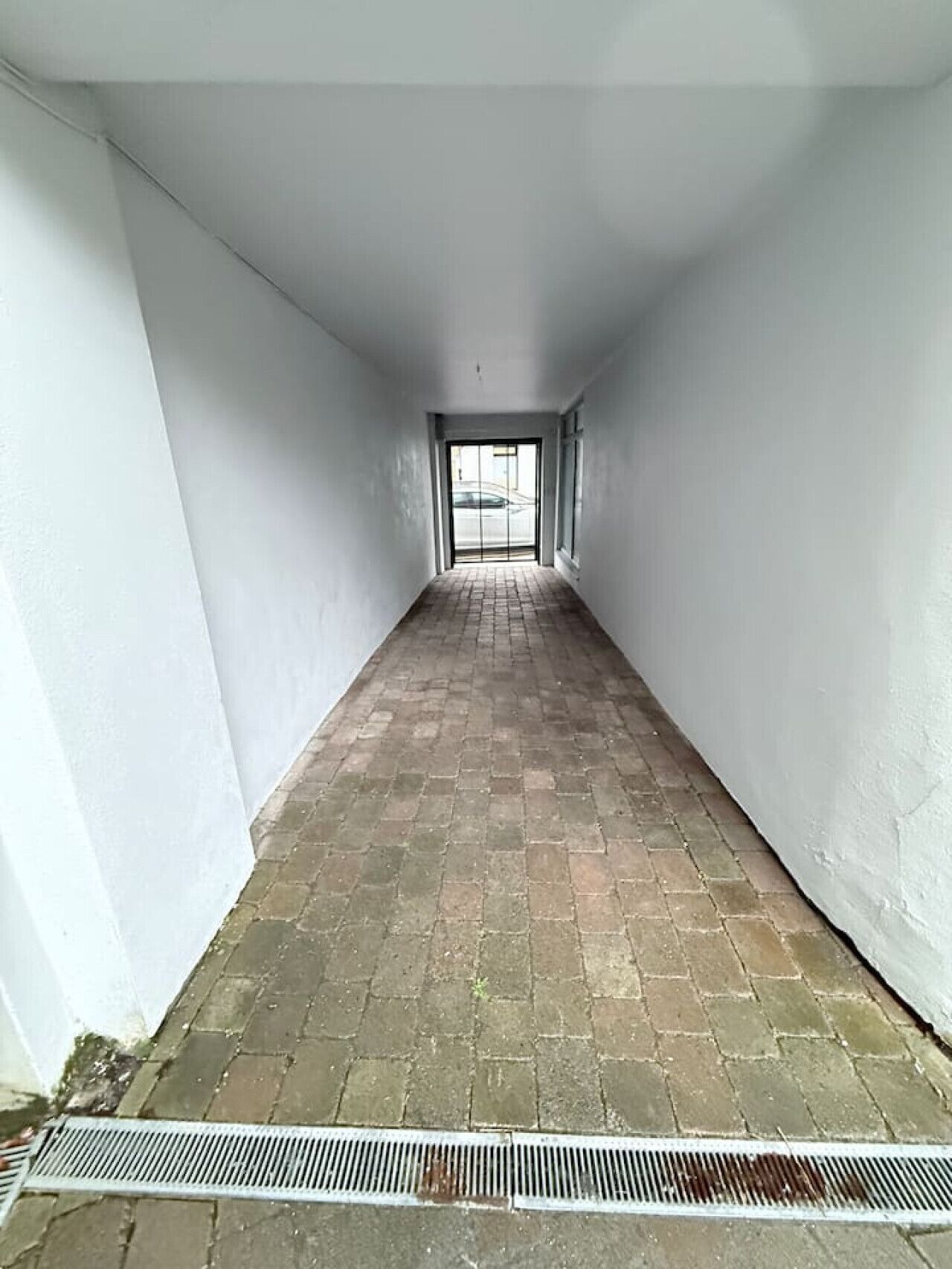Ein schmaler Flur mit weißen Wänden und einem Ziegelboden, der zu einer Tür führt, von der aus ein Auto zu sehen ist.