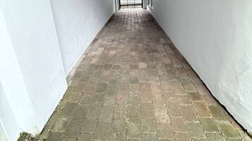 Ein schmaler Flur mit weißen Wänden und einem Ziegelboden, der zu einer Tür führt, von der aus ein Auto zu sehen ist.