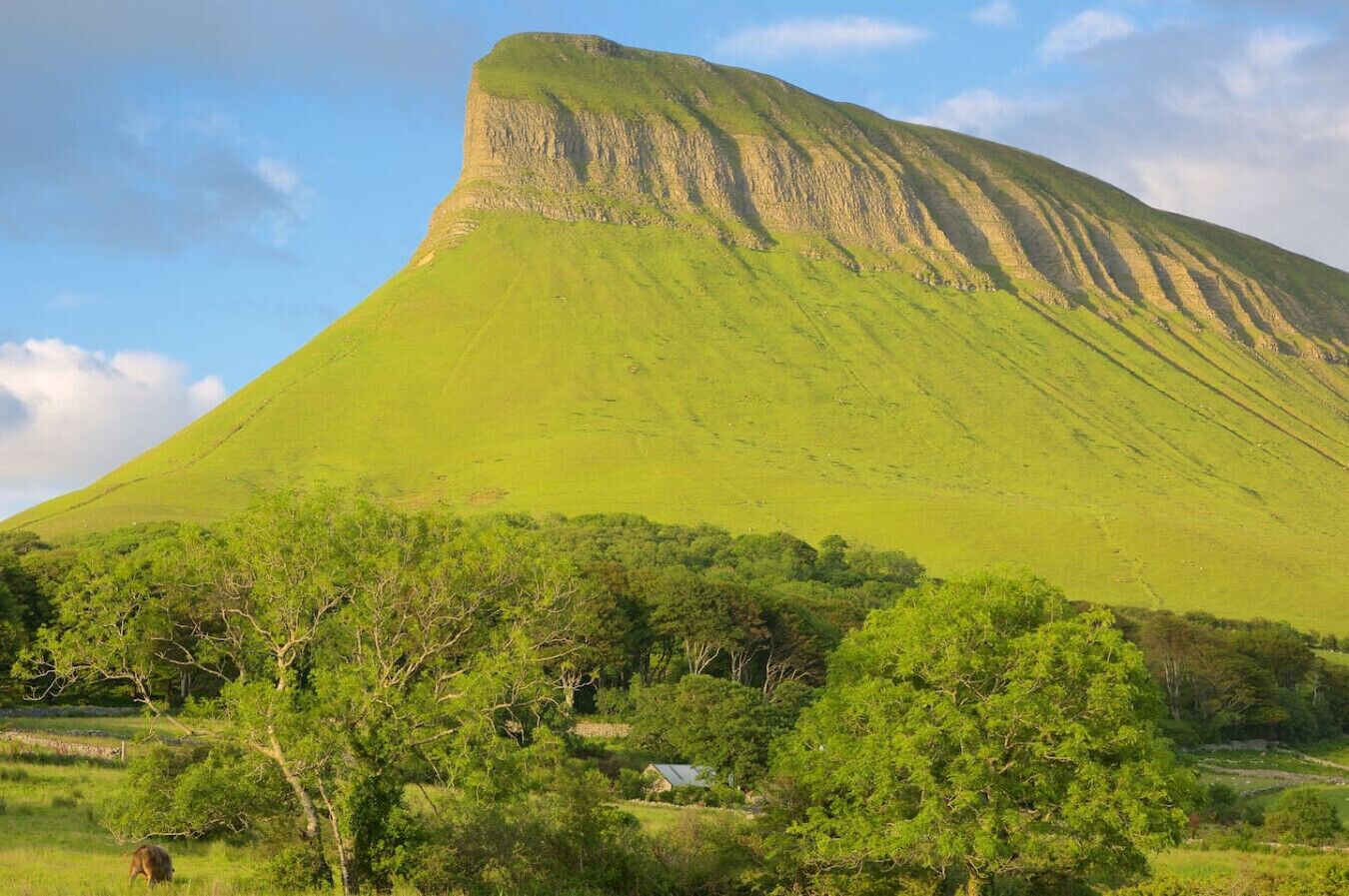 A large, green hill with a flat top and a small house at its base.