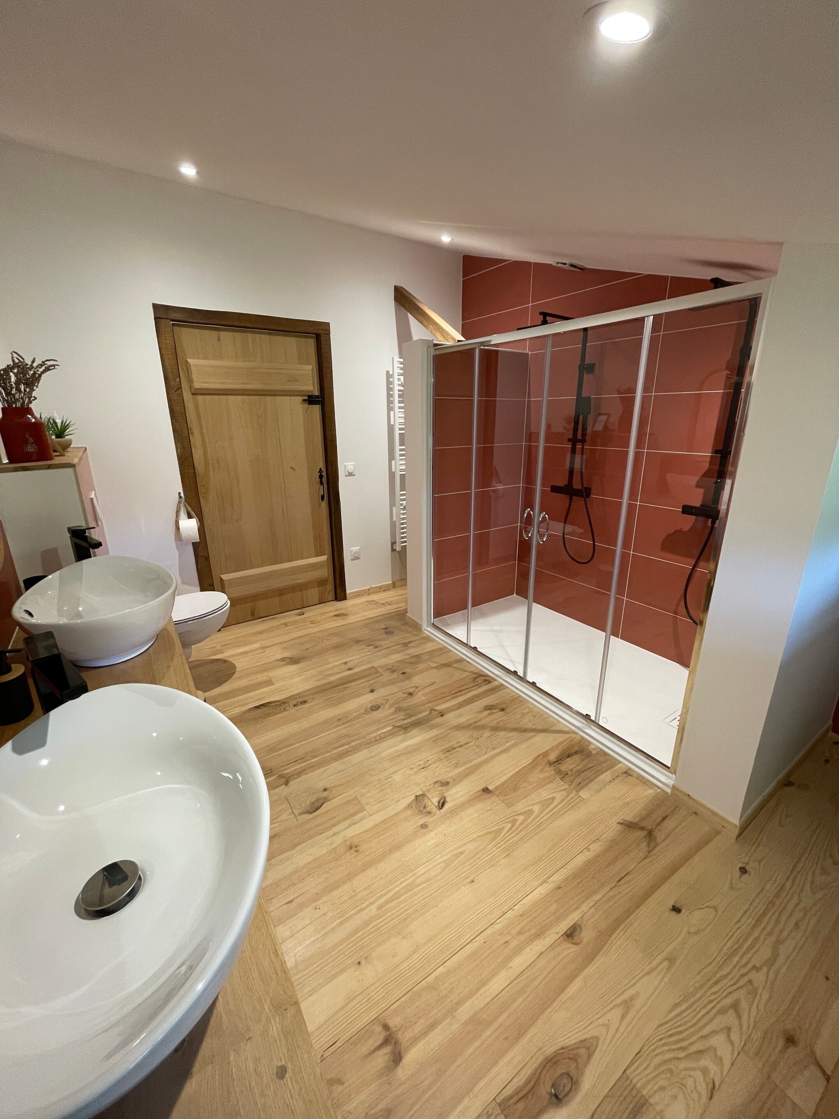 A bathroom with a wooden floor, a white sink, a glass shower enclosure, and a wooden door.