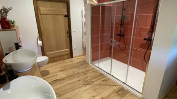 A bathroom with a wooden floor, a white sink, a glass shower enclosure, and a wooden door.