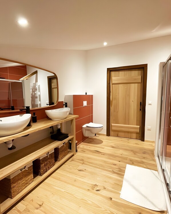 A modern bathroom with a wooden vanity, two sinks, a toilet, and a wooden door.
