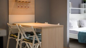 A small, modern room with a wooden desk, a white chair, and a bed with a blue blanket. There is a shelf with bottles and a plant on the wall. - Euro Square Studios (Zagreb)
