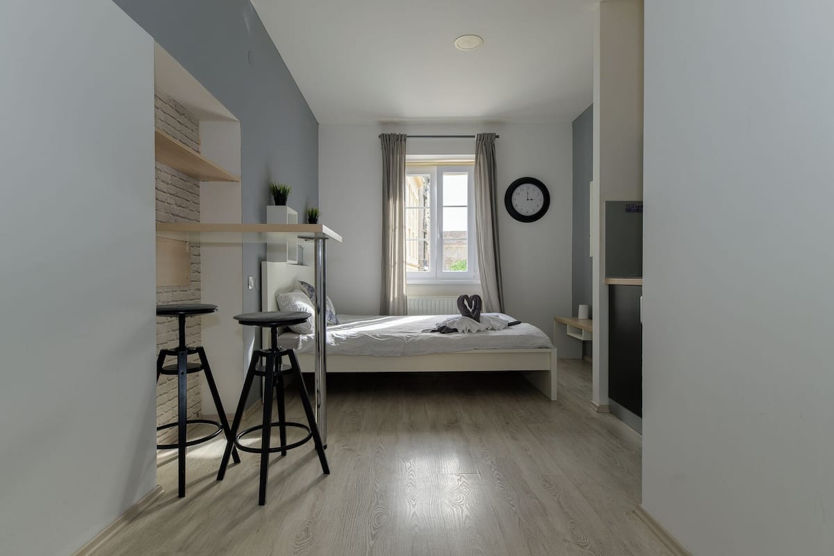 A compact bedroom with a bed, a window with curtains, a wall clock, and a small table with two stools.