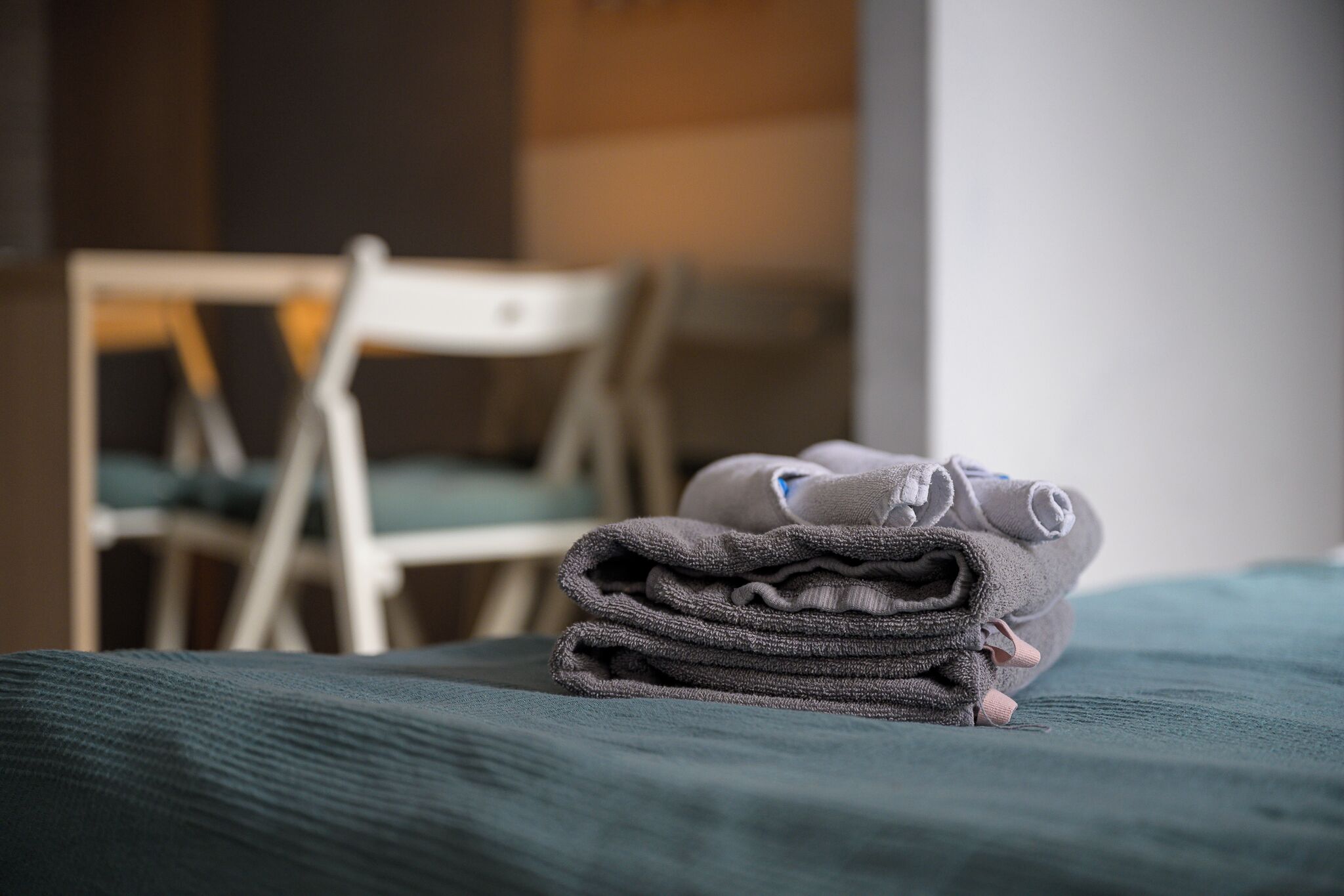Une pile de serviettes soigneusement pliées est posée sur un lit recouvert d’une couverture verte. On aperçoit une chaise blanche à l’arrière-plan et une porte en bois.