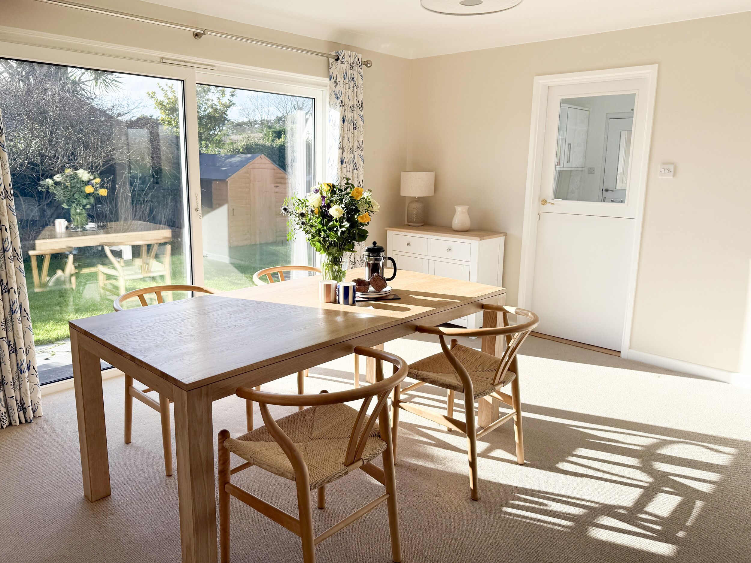 A dining area with a wooden table and chairs, a vase of flowers, and a view of the garden through large windows.
