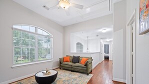 A spacious living room with a large window, a green sofa, a black ottoman, and a patterned rug. - Serenity of Spring, TX & The Woodlands - Modern 4BR Furnished Home (Spring)