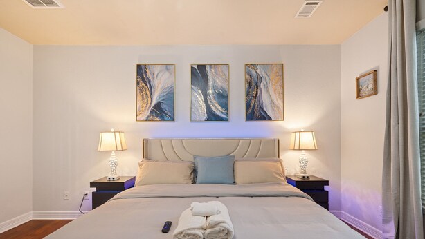 A neatly made bed with a folded towel, two bedside lamps, and three abstract paintings on the wall. - Serenity of Spring, TX & The Woodlands - Modern 4BR Furnished Home (Spring)