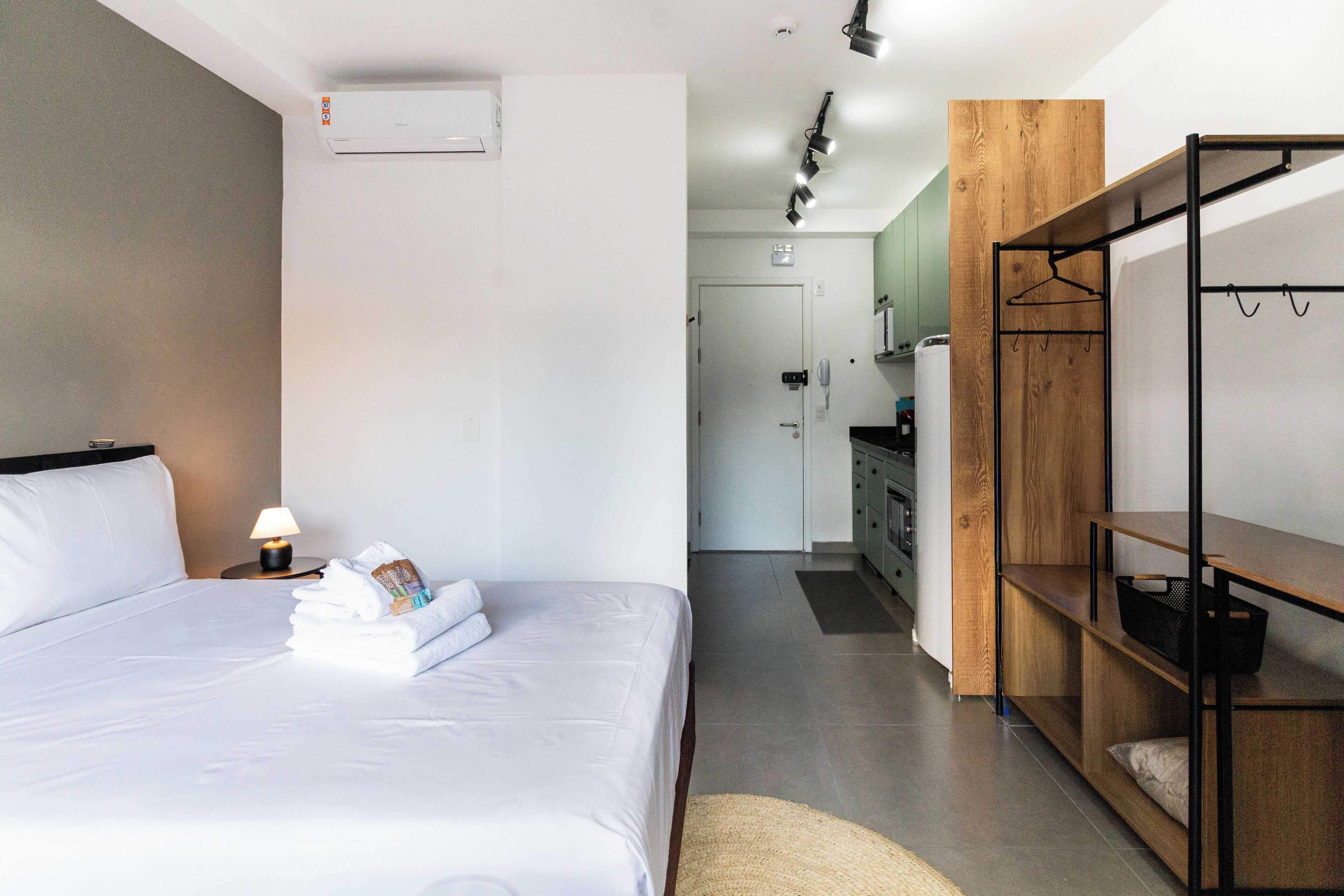A hotel room with a neatly made bed, a bedside lamp, a wooden shelf, and a kitchenette in the background.