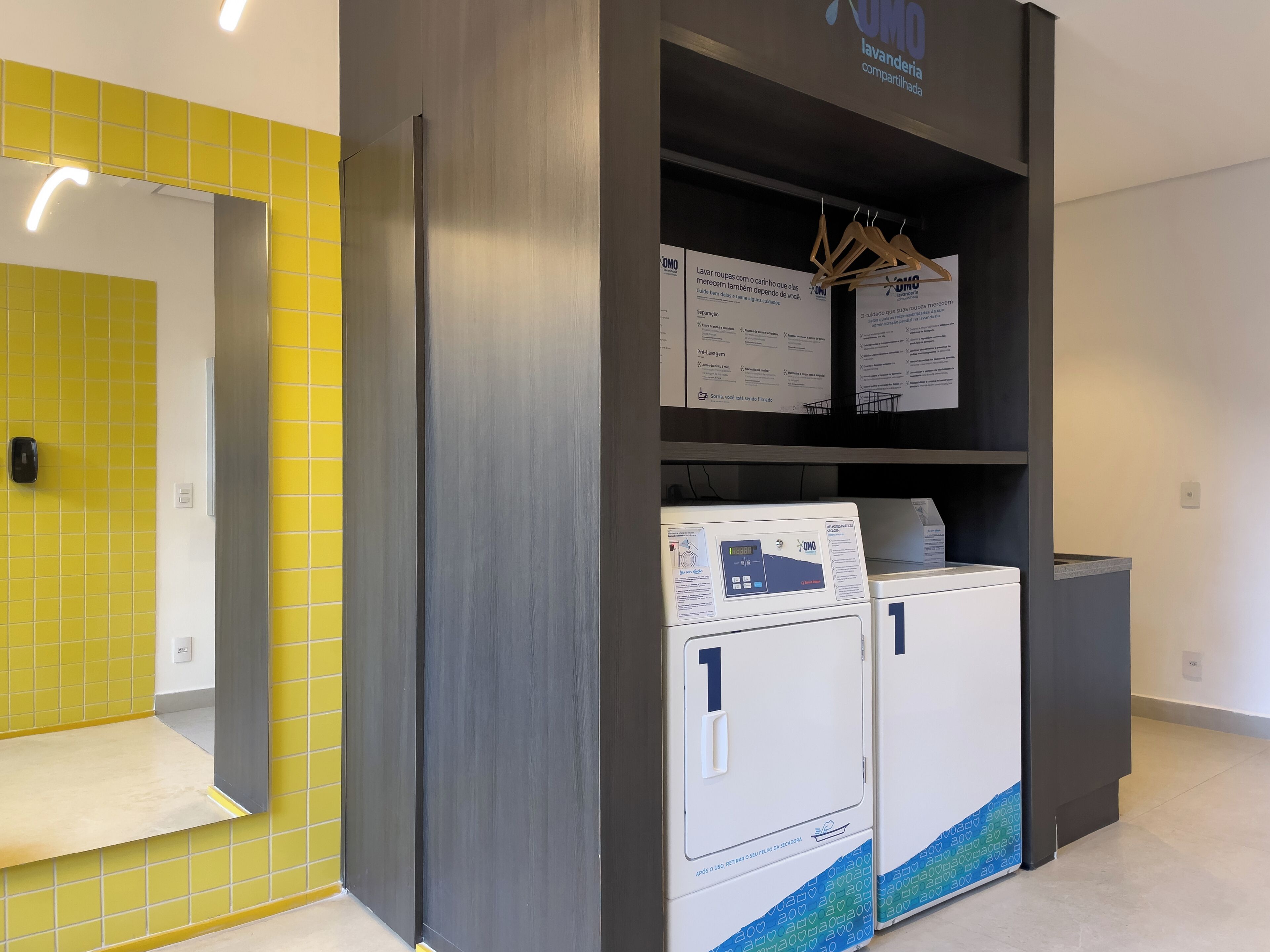 A laundry facility with two front-loading machines, a yellow tiled wall, and informational signs.