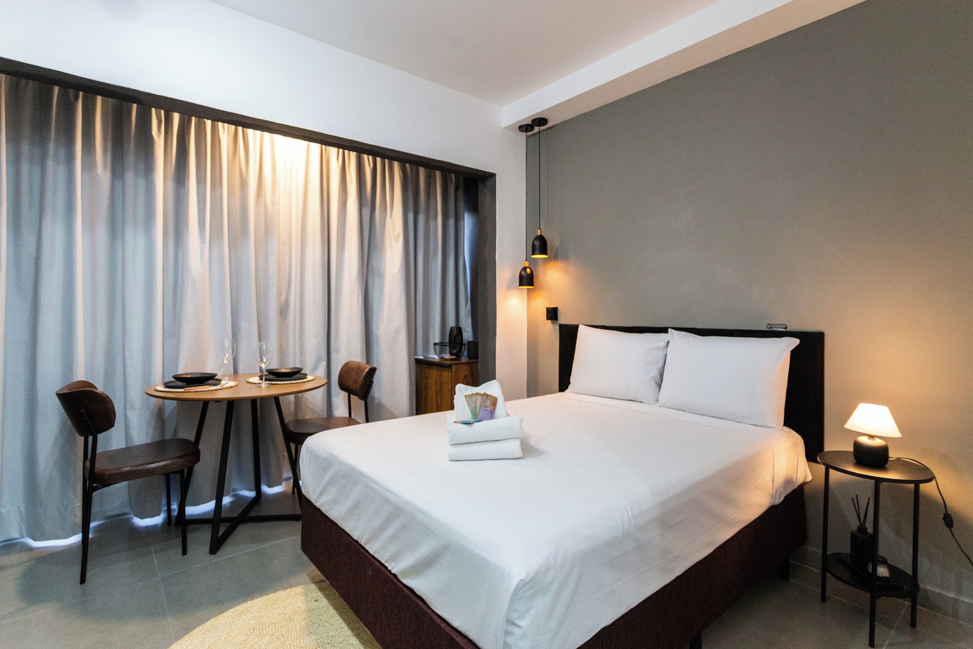 A hotel room with a large bed, a bedside table with a lamp, a small table with chairs, and a window with curtains.