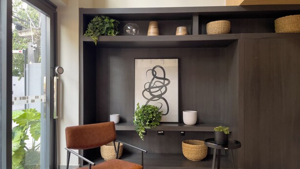 A modern interior with a brown leather chair, a large abstract painting, and a dark wood shelving unit with baskets and decorative items. - La Casa Brooklin (São Paulo)