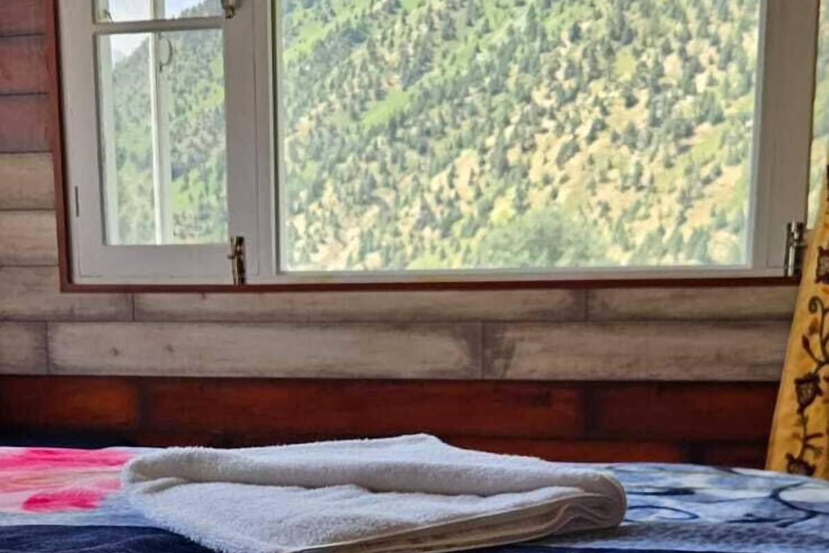 A room with a bed, a window with a view of a forested mountain, and a wooden wall.