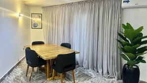 Un coin repas avec une table en bois et des chaises noires, une grande plante en pot et un rideau gris.