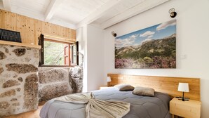 A bedroom with a stone wall, a bed with a grey quilt, a wooden headboard, a nightstand with a lamp, and a window with a view of mountains.