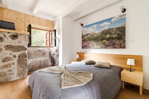 Quarto com parede de pedra, cama com colcha cinza, cabeceira de madeira, mesa de cabeceira com abajur e janela com vista para montanhas.