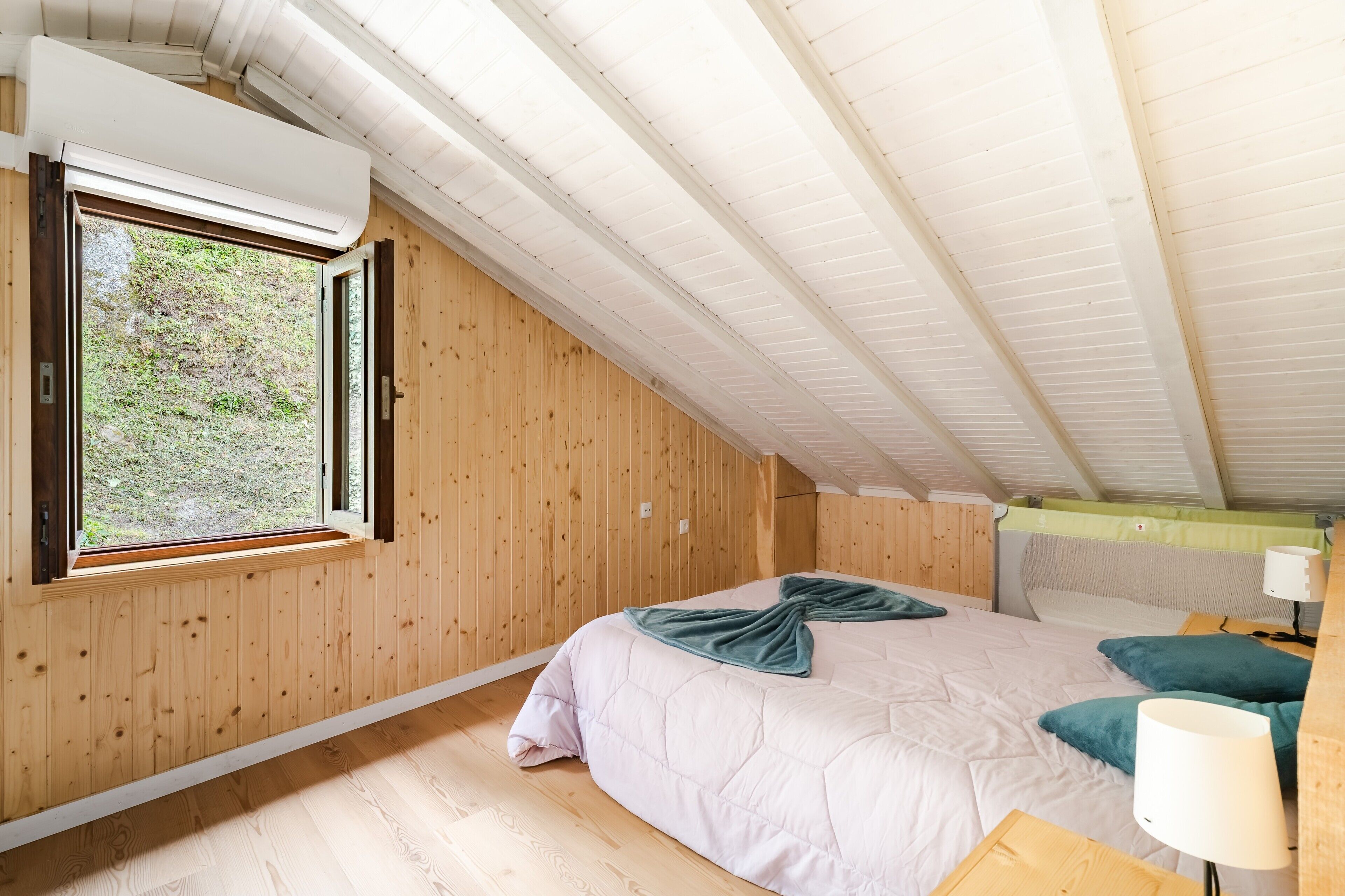 A bedroom with a wooden sloped ceiling, a bed with a quilt, a window, and a bedside table with a lamp.
