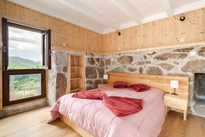 Et soveværelse med stenvæg, træloft, en seng med pink sengetøj, et natbord, et vindue med udsigt og en trækæst.'