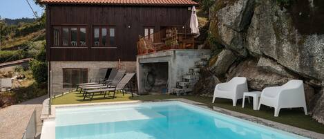 Una piscina con sillas de descanso, un muro de piedra y un edificio de madera con techo rojo.