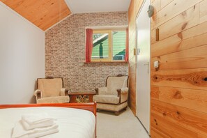 A bedroom with a brick wall, wooden ceiling, a bed, two armchairs, a small table, and a window with red curtains. - 20P 6B Beautiful house in Nature (Scheerwolde)