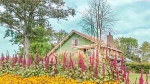 A green house with a porch, surrounded by vibrant flowers and trees. - 20P 6B Beautiful house in Nature (Scheerwolde)