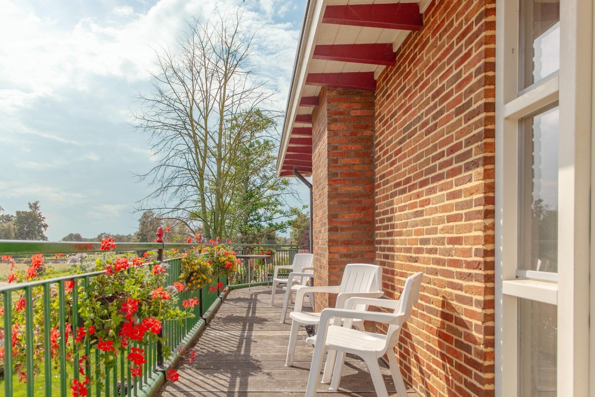 A balcony with white chairs, a brick wall, and a view of trees and a field.
