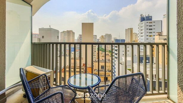 A balcony with a glass table and two wicker chairs, overlooking a cityscape with various buildings. - Modern and Fully Equipped Accommodations Near the Metro and Praca da Republica - Dnrs (São Paulo)