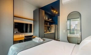 A hotel room with a bed, a TV, a microwave, a refrigerator, a shelf with toiletries, and a large window with a city view. - Modern and Fully Equipped Accommodations Near the Metro and Praca da Republica - Dnrs (São Paulo)