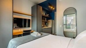 A hotel room with a bed, a TV, a microwave, a refrigerator, a shelf with toiletries, and a large window with a city view. - Modern and Fully Equipped Accommodations Near the Metro and Praca da Republica - Dnrs (São Paulo)