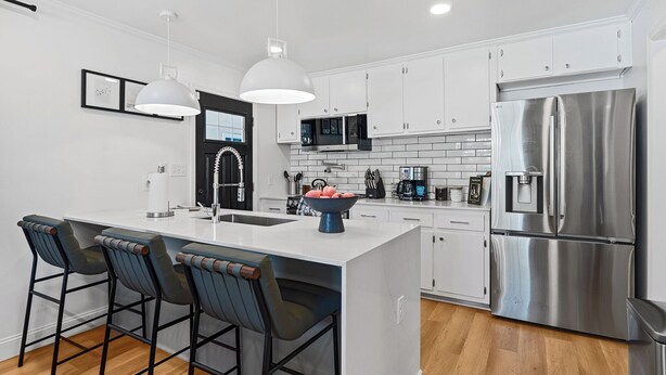 A modern kitchen with white cabinets, a stainless steel refrigerator, a microwave, and a sink. There are three bar stools with dark seats and metal frames.