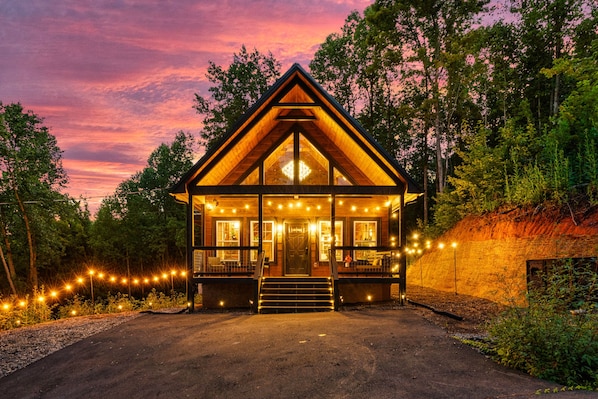 A wooden cabin with a pitched roof, large windows, and a porch with string lights.