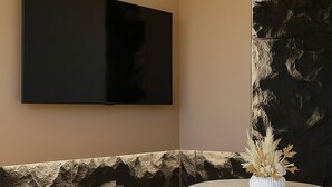 A modern room with a large flat-screen TV mounted on the wall, a round wooden table with a vase of dried flowers, and a textured wall feature. - 3 KING Beds & Free parking • Central Place (Pittsburgh)